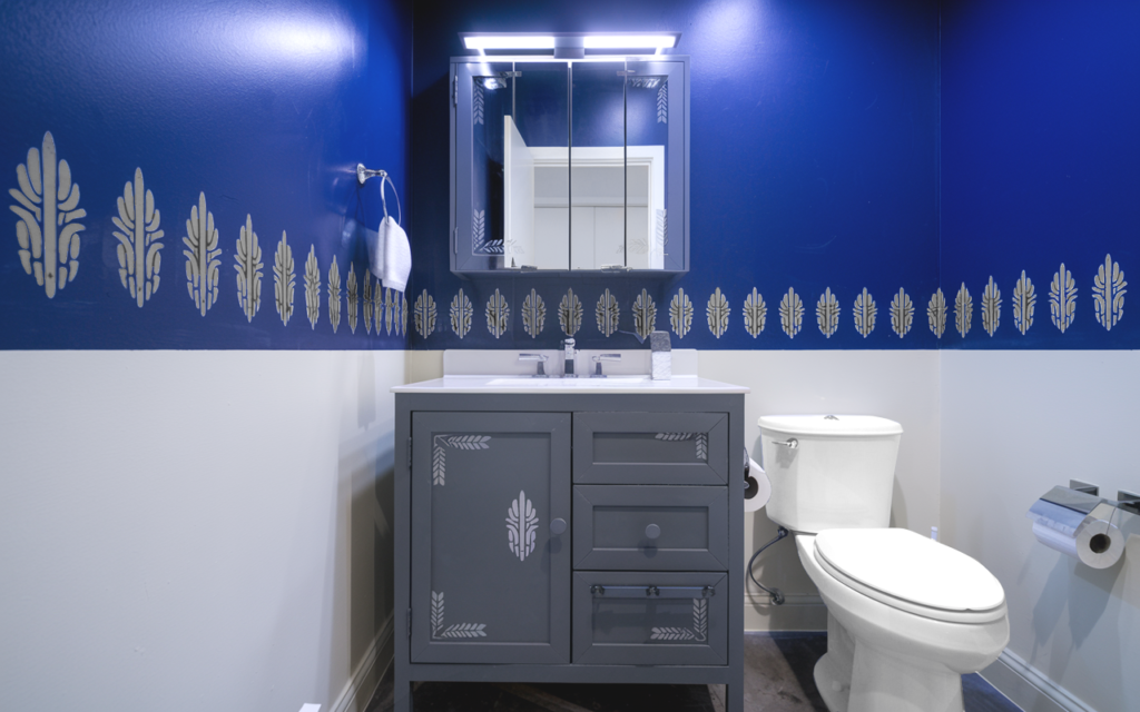 Finished bathroom refresh in Brooklyn, NY featuring two-tone walls with deep blue upper and cream lower sections, cream accents on the blue, a modern grey vanity with lighter grey decorative accents, a white toilet, a glass medicine cabinet, a hanging hand towel on the wall, and a mounted toilet paper holder. Designed by Walls of Style NY