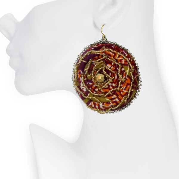 Close-up of a mannequin wearing a handcrafted circular fabric earring with intricate beadwork in gold, bronze, and amber hues. The earring features a swirling pattern with seed beads and metallic accents, creating a bold, artistic statement inspired by African craftsmanship and boho-chic style.